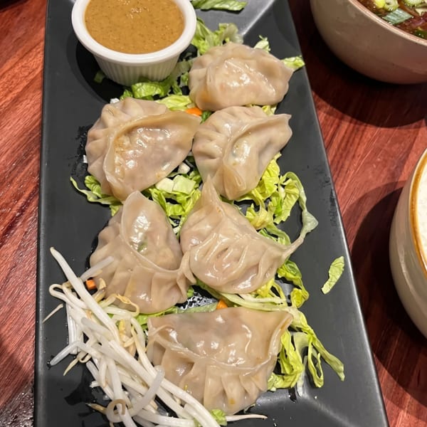 A black rectangular plate of steamed dumplings on lettuce with bean sprouts and a side of peanut sauce.