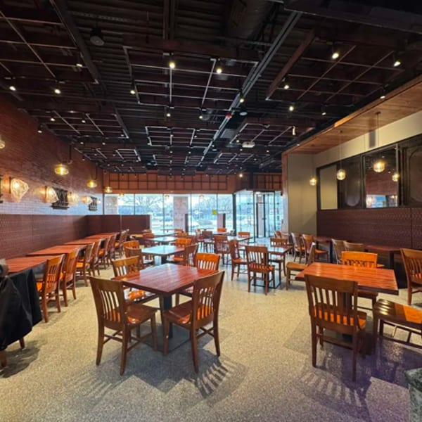 Spacious restaurant interior with numerous empty wooden tables and chairs, brick walls, and large windows.