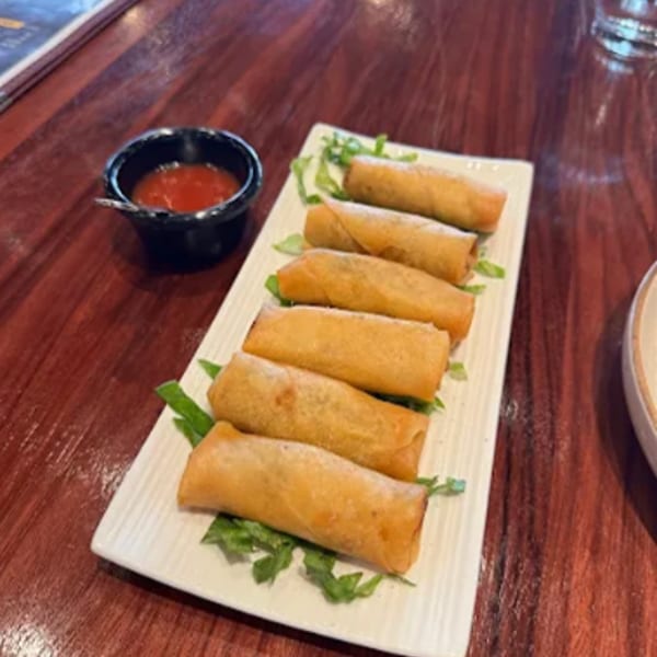 Six crispy spring rolls on a white rectangular plate with a side of red dipping sauce.