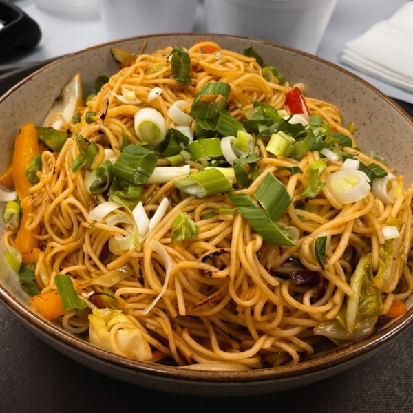 Close-up of a bowl of stir-fried noodles with vegetables and scallions on a dark tray.