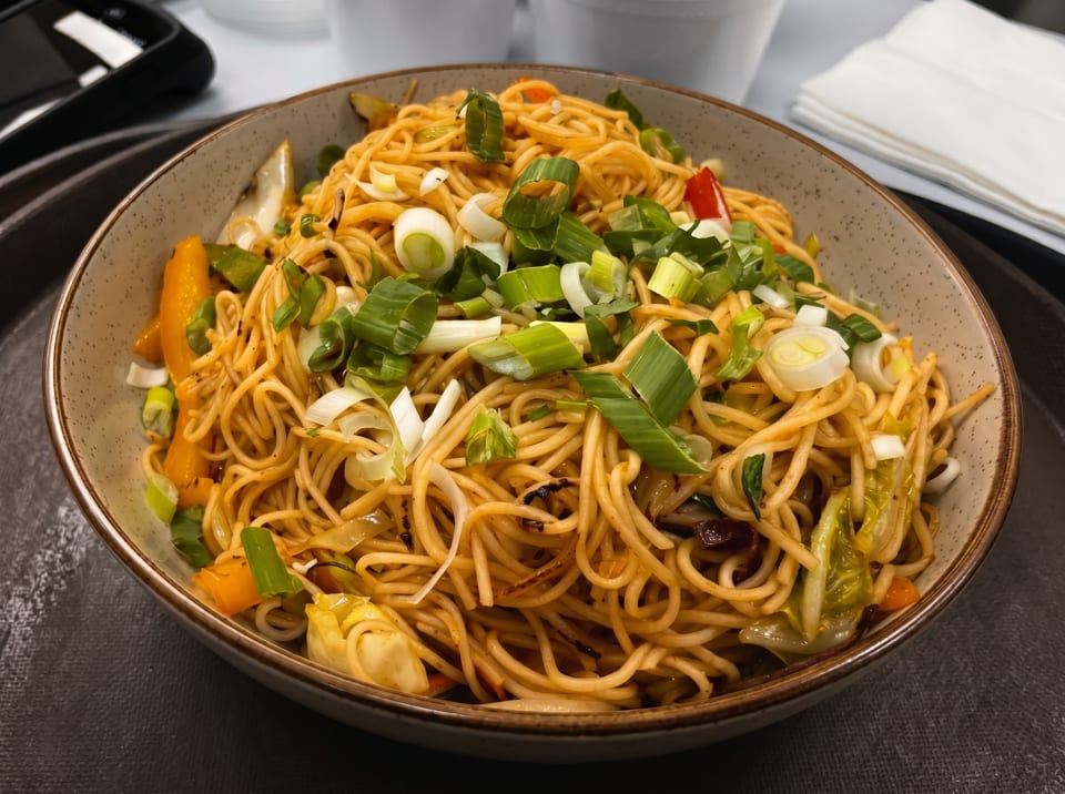 Close-up of a bowl of stir-fried noodles with vegetables and scallions on a dark tray.