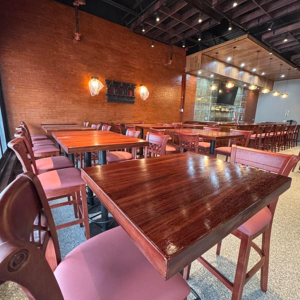 Empty restaurant dining room with dark wooden tables, red chairs, and a brick wall with decorative lighting.