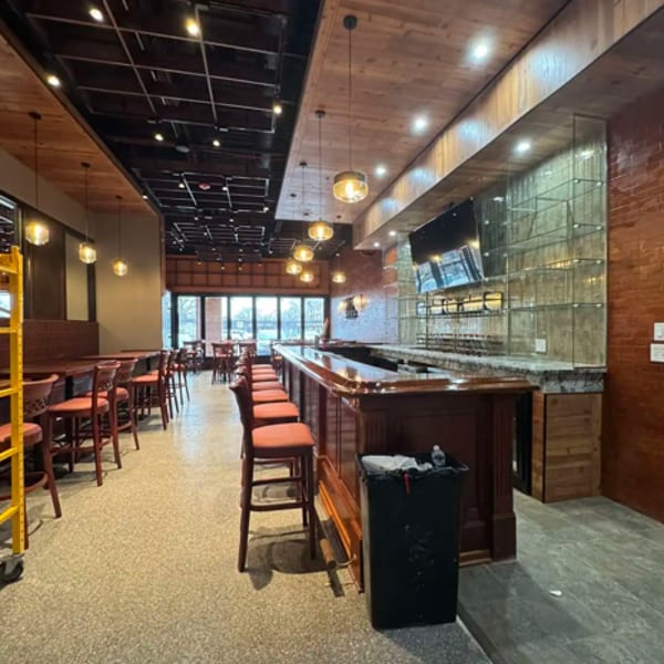 Empty restaurant interior with a long wooden bar, bar stools, and dining tables under warm lighting.