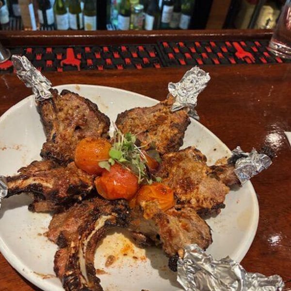 Grilled lamb chops with roasted tomatoes and microgreens on a white plate at a bar.